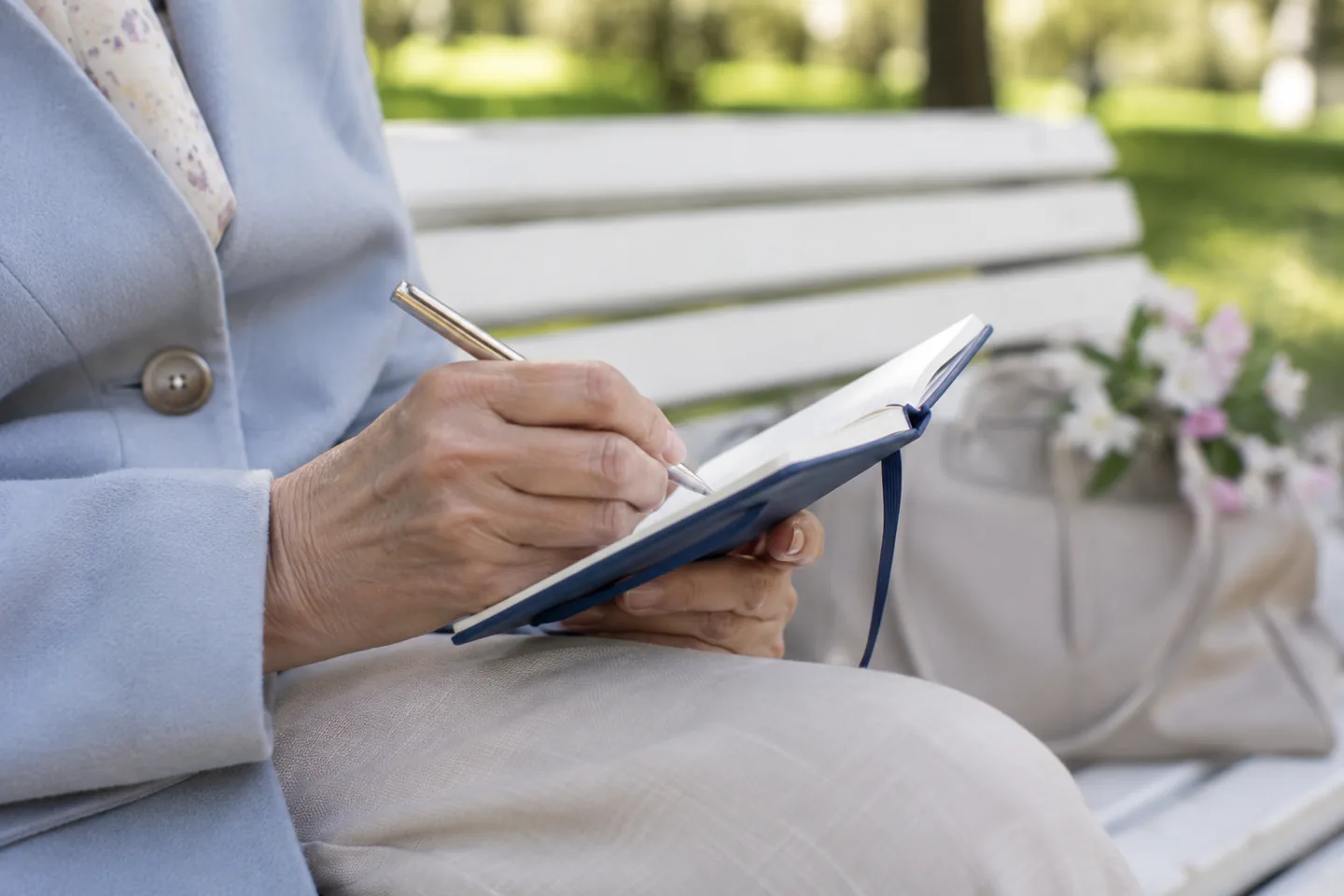 Hands writing in a small blue notebook on a white outdoor bench.