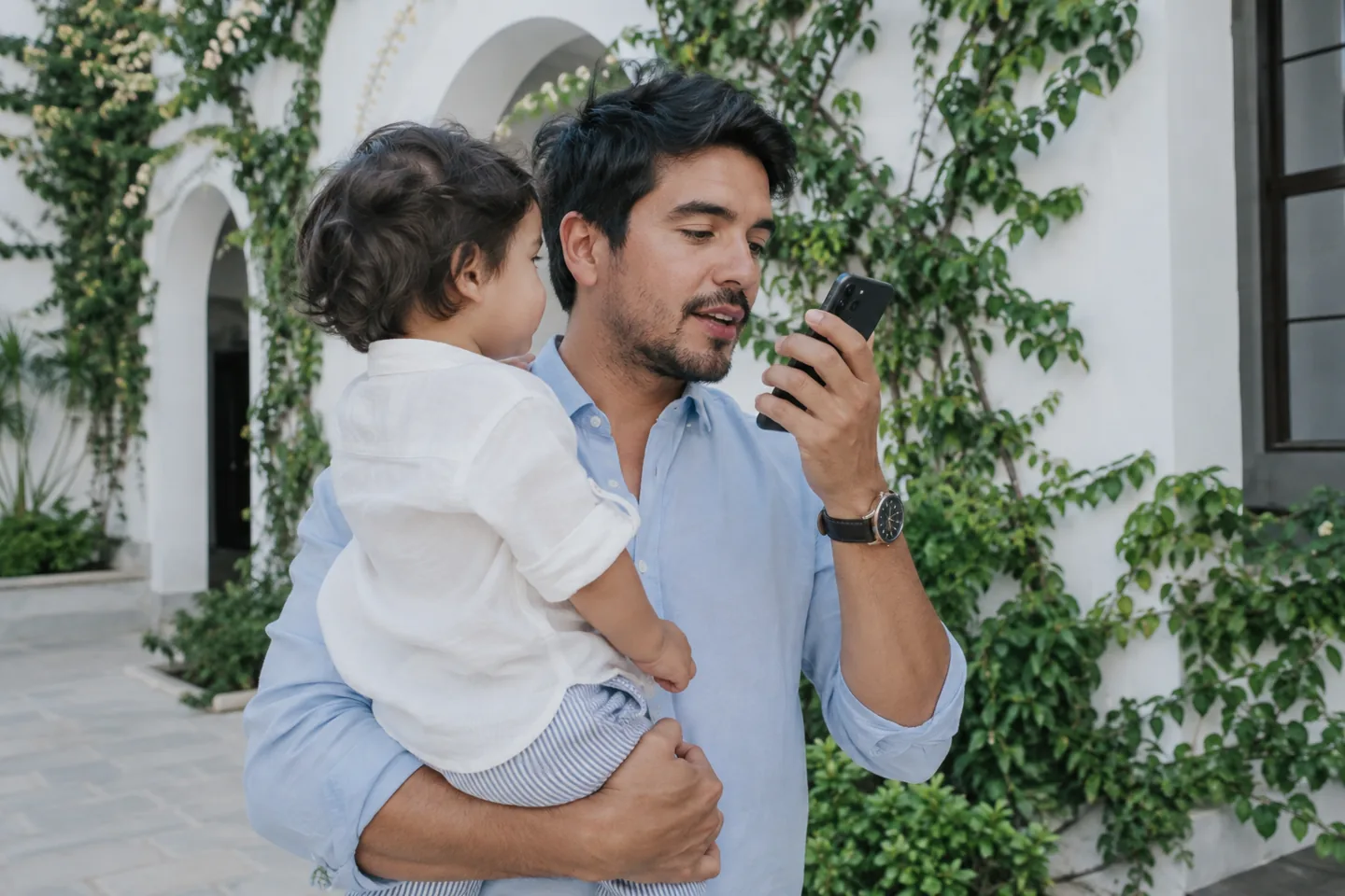 A father holding a child while speaking toward his phone in a courtyard.
